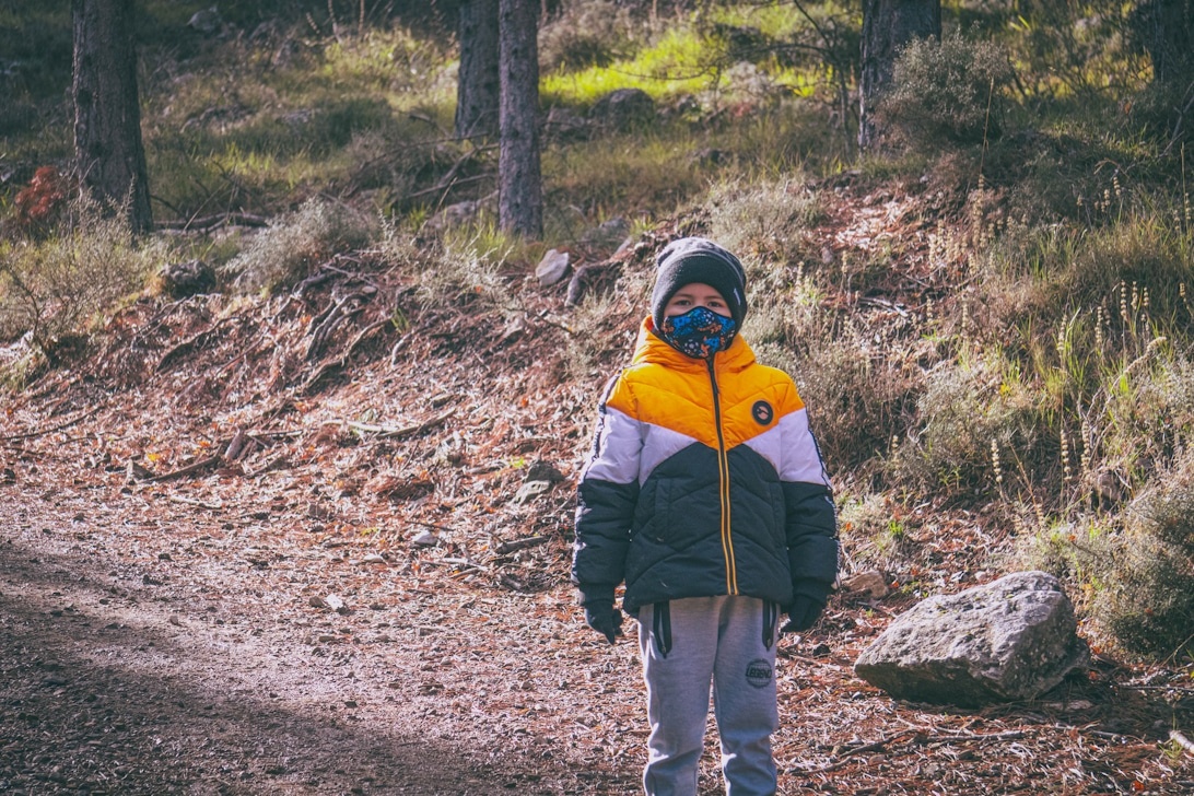 Ein Kind in einer farbenfrohen, mehrlagigen Outdoor-Jacke sitzt in einer Waldlandschaft.
